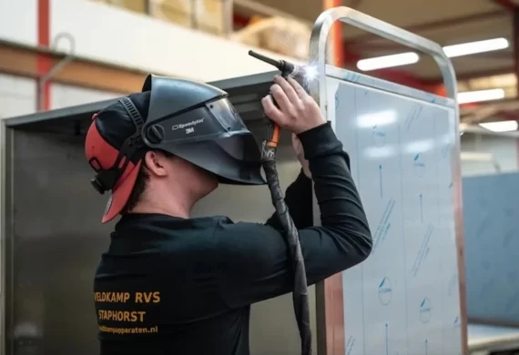 Monteur van Veldkamp RVS in Staphorst die met een lashelm op een RVS constructie last in een werkplaats, waarbij vonken zichtbaar zijn tijdens het precisiewerk.